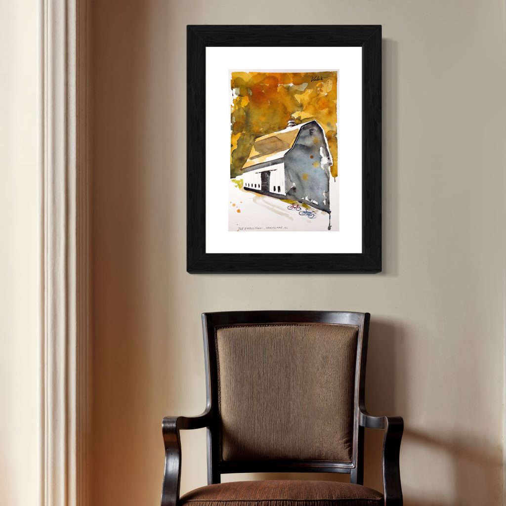 Ewald Barn_framed sq - Michael Valenti Framed abstract watercolor in orange and yellow tones hanging on a beige wall above a dark wood chair.